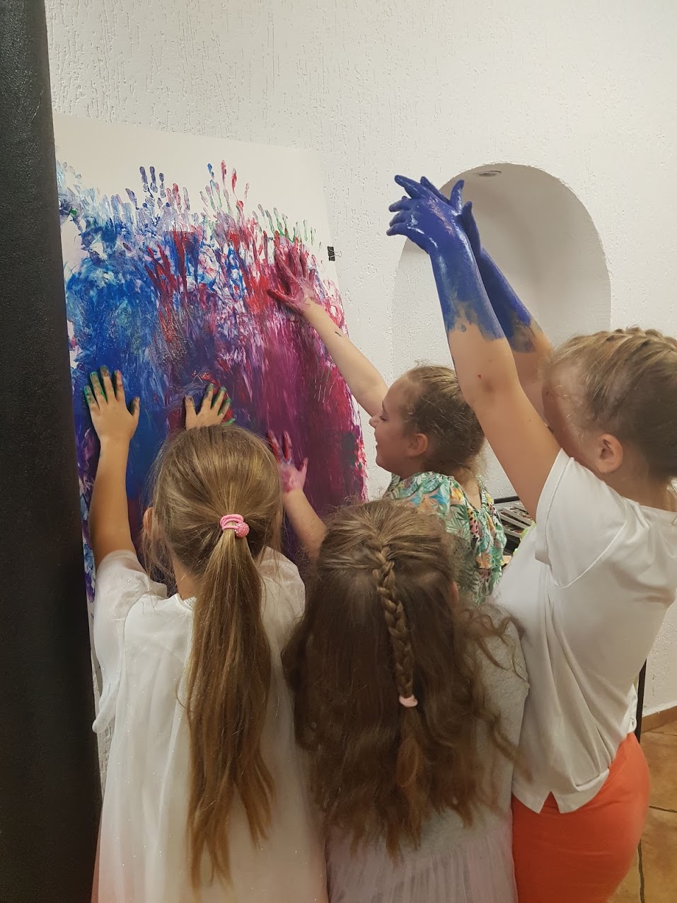 zdjęcie przedstawia dzieci stojące przed sztalugą i malujące rękami umazanymi w kolorowej farbie na białej kartce bez użycia pędzla. Malowanie palcami.